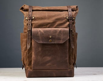 May include: A brown roll-top backpack constructed from waxed canvas and leather. The bag features two vertical leather straps with metal buckles, a large front pocket with a snap closure, and side pockets. The design includes brass-toned hardware.