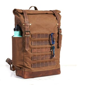 Könnte beinhalten: Ein brauner Rucksack aus gewachstem Segeltuch mit Rollverschluss, Lederriemen mit Metallschnallen, verstärktem Lederboden und einer Seitentasche mit einer türkisfarbenen Trinkflasche. Die Vorderseite hat horizontale Lederriemen und einen blauen Karabiner.