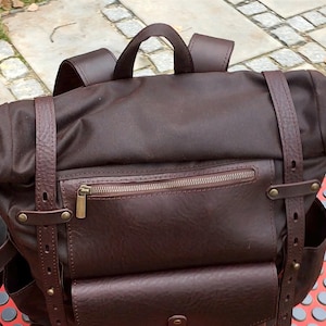 May include: A dark brown roll-top backpack featuring a top handle, two adjustable vertical leather straps with metal rivets, and a horizontal zipped front pocket. The bag includes side pockets and a flap-covered lower front compartment.