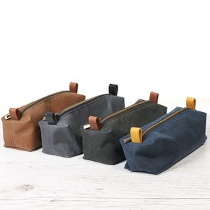 Op de afbeelding: Vier rechthoekige etuis met ritssluiting op een rij. De etuis zijn gemaakt van een matte, gestructureerde stof in de kleuren bruin, grijs, donkergroen en marineblauw. Elk etui is voorzien van een metalen ritssluiting en een klein lipje.