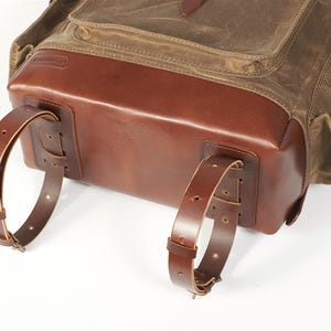 May include: A backpack featuring a dark olive green waxed canvas upper section and a smooth, polished brown leather base. Two adjustable brown leather straps are attached to the bottom, each secured with metal buckles and multiple adjustment holes.