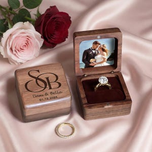 Könnte beinhalten: Eine Ringbox aus dunklem Holz mit Klappdeckel, graviert mit den Buchstaben SB, den Namen Sam & Bella und dem Datum 01.12.2025. Die geöffnete Box enthält einen Goldring mit einem großen runden Stein und ein Foto eines Paares im Deckel.