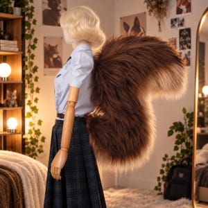 Op de afbeelding: Een etalagepop met blond haar draagt een lichtblauw shirt met korte mouwen en een donkergrijze geruite rok, voorzien van een grote, pluizige, bruin en crèmekleurige imitatiebontstaart aan de achterkant, die in een gebogen vorm naar buiten uitsteekt.