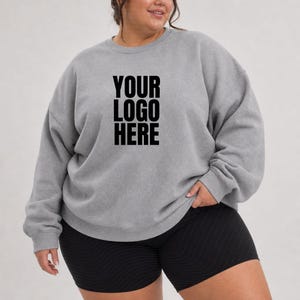May include: A heather grey crewneck sweatshirt featuring the black text YOUR LOGO HERE printed on the centre chest. The garment has long sleeves with ribbed cuffs and a ribbed neckline, paired with solid black athletic shorts.