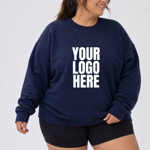 Può includere: Una felpa blu navy a maniche lunghe con girocollo, caratterizzata dalla scritta bianca YOUR LOGO HERE stampata in un carattere sans-serif in grassetto al centro del petto. Il capo presenta scollo, polsini e orlo a coste.