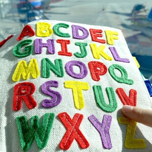 May include: A white fabric book with the alphabet embroidered in colourful felt letters. The letters are in different colours, including red, yellow, green, purple, and blue. The book is being held by a hand.
