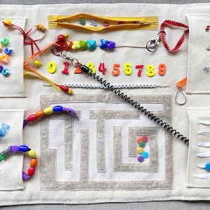 May include: A beige fabric activity mat with various sensory elements, including a zipper, buttons, beads, a maze, and a coil. The mat also features colorful numbers, letters, and a rainbow-colored string.