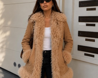 May include: A tan suede coat featuring a long, curly, shaggy faux fur trim along the wide lapels, cuffs, and hemline. The coat is worn over a plain white tank top and black denim jeans, paired with dark-tinted oval sunglasses and small hoop earrings.