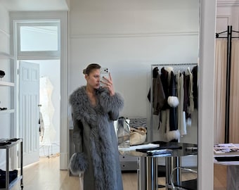 May include: A long, grey leather coat featuring a thick, fluffy grey fur trim along the wide collar, the front opening, the sleeve cuffs, and the bottom hem. The coat has a smooth, matte finish and reaches down to the lower calf area.