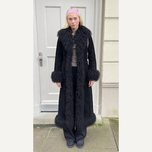 May include: A long black coat featuring a textured black fur trim along the collar, front opening, cuffs, and hem. The coat is worn over a brown top and black trousers, paired with a light pink headband, creating a layered look with distinct textures.