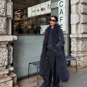 May include: A long, dark charcoal gray coat featuring a thick, textured faux fur trim along the lapels, cuffs, and hem. The coat is worn over a dark gray sweater and loose-fitting black trousers, paired with black shoes and dark oval sunglasses.