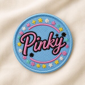 May include: A circular embroidered patch with a light blue background and a pink border. The word Pinky is stitched in large pink letters with a black outline. Small, colorful embroidered flowers and stars surround the central text.