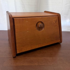 May include: A rectangular wooden bread box with a medium brown finish. The box features a front-hinged door that opens downward, equipped with a small, circular wooden knob centered on the panel. The sides have rounded edges and a smooth texture.