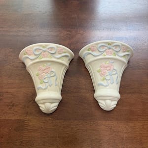 May include: A pair of cream-colored ceramic wall pockets featuring raised relief designs of pink roses, green leaves, and light blue ribbons. Each piece has a flared top edge and a tapered base with a molded fan-like detail at the bottom.
