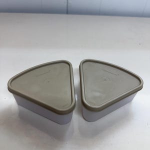 May include: Two triangular food storage containers with white bases and tan lids. Each lid features the Tupperware brand name embossed in the center. The containers have rounded corners and a flat top surface, designed for stacking or organizing food items.