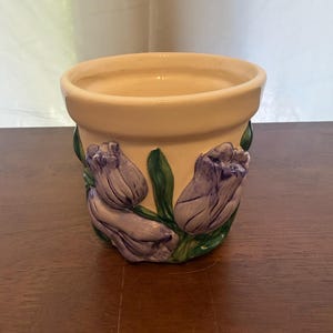 May include: A cream-colored ceramic flower pot featuring a raised, three-dimensional relief design of purple tulips with green stems and leaves wrapped around the exterior. The pot has a wide, smooth rim and a cylindrical shape with a glossy finish.