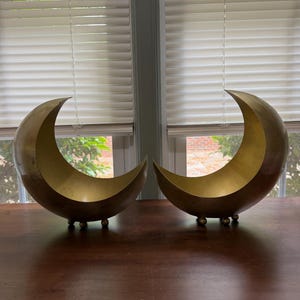 May include: Two crescent moon-shaped bowls crafted from polished brass. Each bowl features a smooth, curved interior and exterior surface with a metallic gold finish, supported by three small, spherical ball feet at the base.