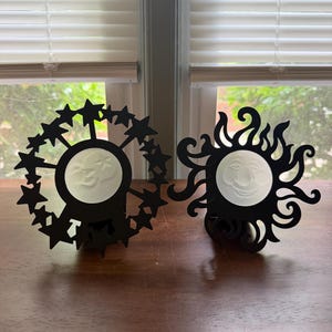 May include: Two black metal decorative stands featuring circular frosted glass inserts. One stand is surrounded by a ring of stars with a moon face insert, while the other features swirling sun rays surrounding a sun face insert.