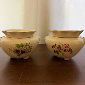May include: Two cream-colored ceramic bowls with scalloped lids and three small feet. The left bowl features a floral pattern with purple and white blossoms, while the right bowl displays a design of hummingbirds and pink flowers.