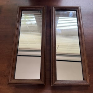 May include: Two rectangular mirrors with dark brown wooden frames. Each mirror features a simple, clean-lined border design. The glass surfaces are clear and reflective, showing a view of horizontal window blinds in the reflection.