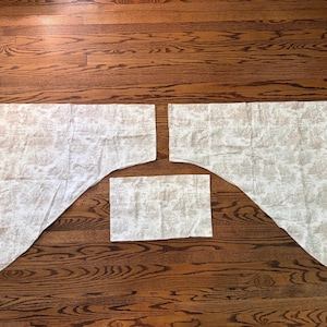 May include: Three pieces of fabric with a light beige and white toile pattern. Two pieces are rectangular with one diagonal edge forming a point, and the third piece is a smaller, simple rectangle. The fabric features a repeating landscape scene.