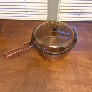 May include: A round, amber-colored glass saucepan with a matching transparent amber glass lid. The pan features a long, integrated glass handle extending from one side and a small, circular knob handle centered on the top of the lid.