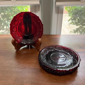May include: A set of deep red glass saucers featuring a pressed pattern with a scalloped edge and a central sunburst design. One saucer is displayed upright on a wooden stand, while a stack of matching saucers rests flat nearby.