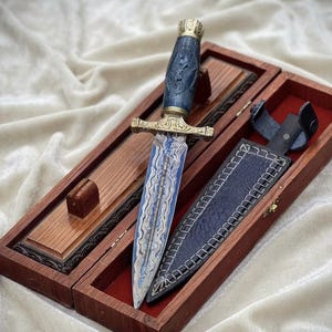 Könnte beinhalten: Ein Dolch mit einer blau-silbern gemusterten Damaststahlklinge, einem Parierstange aus Messing und einem dunkelblauen Griff mit Schnitzereien. Er wird in einer rechteckigen Holzschatulle mit rotem Innenfutter präsentiert, inklusive einer dunkelblauen Scheide.