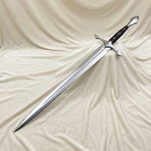 May include: A double-edged steel sword featuring a pointed blade, a silver-toned metal crossguard, and a dark brown wooden handle with a silver-toned pommel. The blade has a central fuller running down the length of the metal surface.