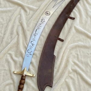 Peut inclure: Un sabre courbe à pointe fendue, orné d'une calligraphie arabe gravée sur la lame et d'un emblème circulaire. La poignée est en bois sombre avec une garde et un pommeau en laiton. Un fourreau en cuir marron assorti avec des lanières est posé à côté.