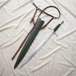 May include: A steel sword with a dark green grip and a crossguard, accompanied by a matching dark green scabbard. The scabbard is attached to a brown leather harness system featuring adjustable straps, metal buckles, and circular metal rings.