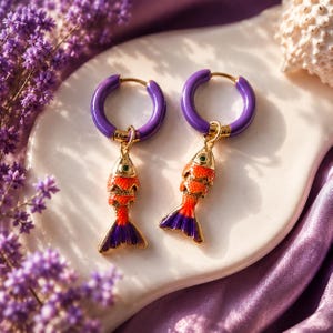 Op de afbeelding: Een paar oorringen met paarse emaille hoepels en bungelende visbedels. De visbedels zijn gekleurd in oranje en rood met paarse staarten en kleine groene ogen. De oorbellen zijn afgewerkt met goudkleurige metalen accenten en een stevige sluiting.