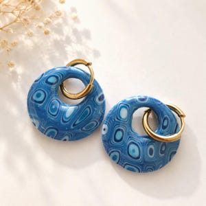 Op de afbeelding: Een paar oorringen met ronde blauwe hangers met een gemarmerd patroon van licht- en donkerblauwe wervelingen. Elke hanger hangt aan een dunne, gepolijste goudkleurige metalen ring, wat een samenhangende set sieraden vormt.