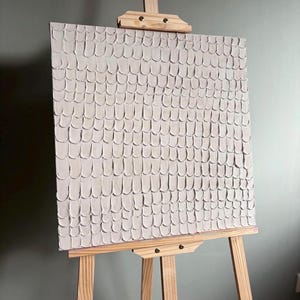 May include: A square canvas painting featuring a light beige, monochromatic impasto texture. The surface is covered in a repeating pattern of small, rounded, overlapping brushstrokes that create a raised, tactile effect across the entire piece.