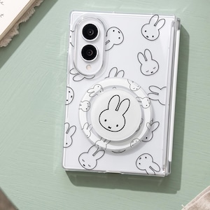 Op de afbeelding: Een doorzichtig smartphonehoesje met een herhalend zwart lijnpatroon van het konijn Miffy. Het hoesje bevat een ronde witte telefoonhouder aan de achterkant, gecentreerd over het patroon, met daarop een zwart lijntekening van Miffy.