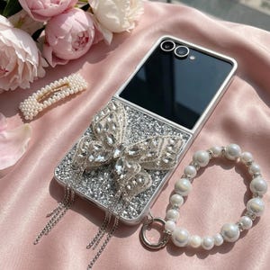 May include: A foldable smartphone case featuring a silver glitter surface, a large crystal-encrusted butterfly ornament, and dangling silver chains. A matching white pearl beaded wrist strap with silver accents is attached to the side of the case.