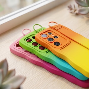 May include: Three smartphone cases stacked together, featuring a vibrant gradient design in pink, lime green, and orange. Each case includes a raised camera lens protector and an integrated circular finger ring strap attached to the top edge.