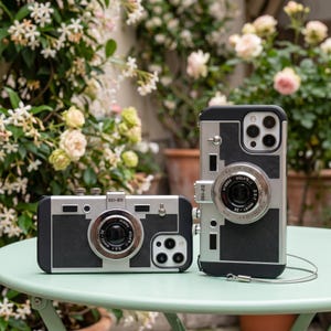 Puede incluir: Dos fundas para teléfono negras y plateadas diseñadas para parecer cámaras vintage. Cada funda presenta una protuberancia de lente circular central, paneles negros texturizados y detalles metálicos plateados. El texto D2-100 y D2-120 está impreso.