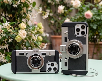 May include: Two black and silver phone cases designed to resemble vintage cameras. Each case features a central circular lens protrusion, textured black panels, and silver metallic accents. The text D2-100 and D2-120 is printed on the cases.