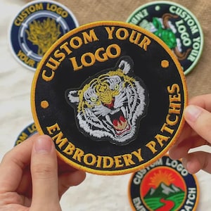 Peut inclure: Un écusson brodé circulaire présentant une tête de tigre centrale avec une fourrure blanche, noire et jaune et une gueule ouverte. La bordure noire contient le texte brodé jaune CUSTOM YOUR LOGO EMBROIDERY PATCHES avec deux petits points jaunes.