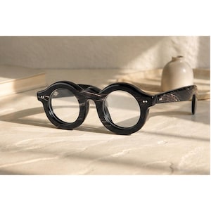 Könnte beinhalten: Runde Brille mit einem dicken, schwarzen Rahmen mit einer subtilen Holzmaserung. Die Brille hat klare Gläser, einen markanten Steg und zwei kleine silberfarbene Nieten auf jeder Seite des vorderen Rahmens, was ein kühnes, kreisförmiges Design ergibt.