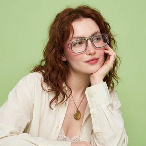 May include: Spectacles featuring a double bridge design with a clear, translucent pink frame and a contrasting dark green inner rim. The glasses have a rectangular shape with rounded corners and clear lenses, providing a distinct layered colour effect.