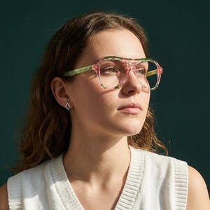 Könnte beinhalten: Brille im Fliegerstil mit einem transparenten rosa Rahmen und grünen Marmor-Akzenten entlang des Stegs und der Bügel. Die Brille verfügt über ein Doppelsteg-Design und klare Gläser, was ihr ein markantes Aussehen mit heller Rahmenstruktur verleiht.
