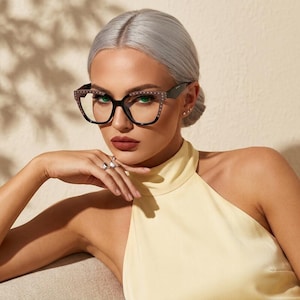 May include: Oversized cat-eye glasses featuring a black frame with a subtle studded pattern along the upper rim and temples. The glasses have clear lenses and a distinct angular shape that frames the face, providing a bold and defined look.