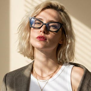 Może przedstawiać: Czarne okulary typu kocie oczy z grubymi oprawkami i przezroczystymi szkłami. Okulary mają ostry, kanciasty kształt. Są noszone z białym prążkowanym topem na ramiączkach, brązową marynarką o wyraźnej fakturze oraz wielowarstwowym złotym naszyjnikiem.