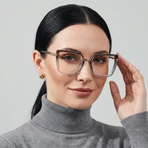May include: Oversized square spectacles featuring a translucent grey gradient frame with a subtle ombre effect. The glasses have thin metallic gold-tone temples and a bridge, providing a lightweight look with clear lenses and a modern geometric shape.