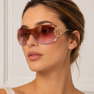 May include: Rimless sunglasses featuring a single-piece gradient lens that transitions from a deep brown to a light pink hue. The gold-tone metal temples are adorned with a decorative coiled snake motif embellished with small clear crystals.
