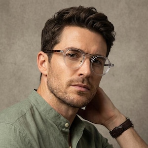 May include: Round spectacles with clear frames and a tortoiseshell brow bar detail. The glasses feature a thin bridge and slim temples, paired with a sage green button-down shirt and a dark brown leather watch on the wrist.