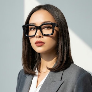 Può includere: Occhiali con montatura nera spessa e forma quadrata. Gli occhiali presentano una finitura lucida e un ponte che collega le due lenti. Le montature sono realizzate con un design audace e prominente che poggia sul ponte del naso.