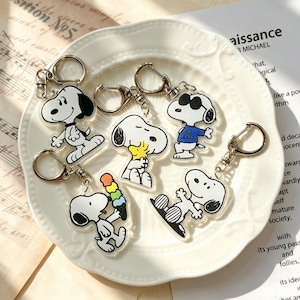 Peut inclure: Cinq porte-clés en acrylique transparent représentant Snoopy dans diverses poses, dont un portant des lunettes de soleil et un t-shirt bleu marqué Joe Cool, un tenant une glace colorée, un serrant Woodstock, et un debout, avec des fermoirs en métal argenté.